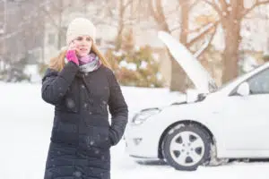 Image of woman using a smartphone to call for roadside assistance. A broken down car is shown with the hood up in the wintry background.
